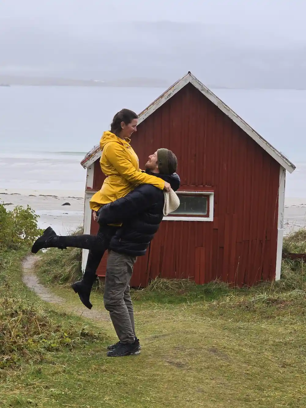Person in yellow jacket lifted by another near red coastal hut