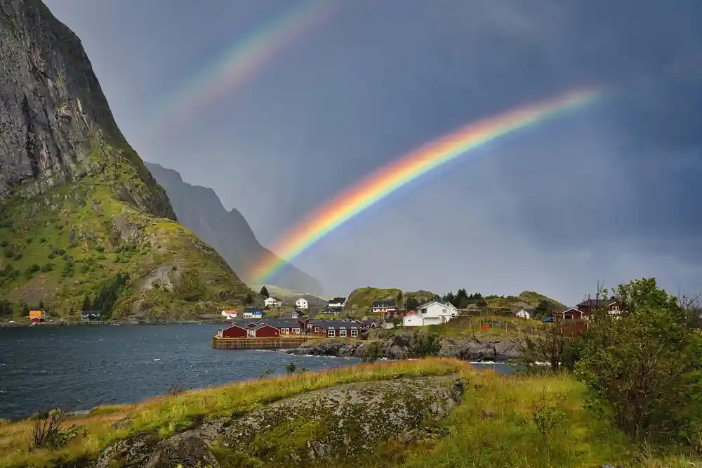 Rainbow over red fishing huts beside water and green hillside