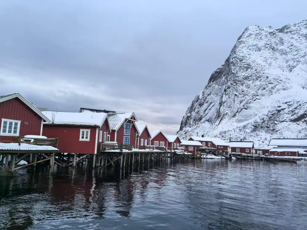 Red wooden buildings on stilts below steep snowy cliff