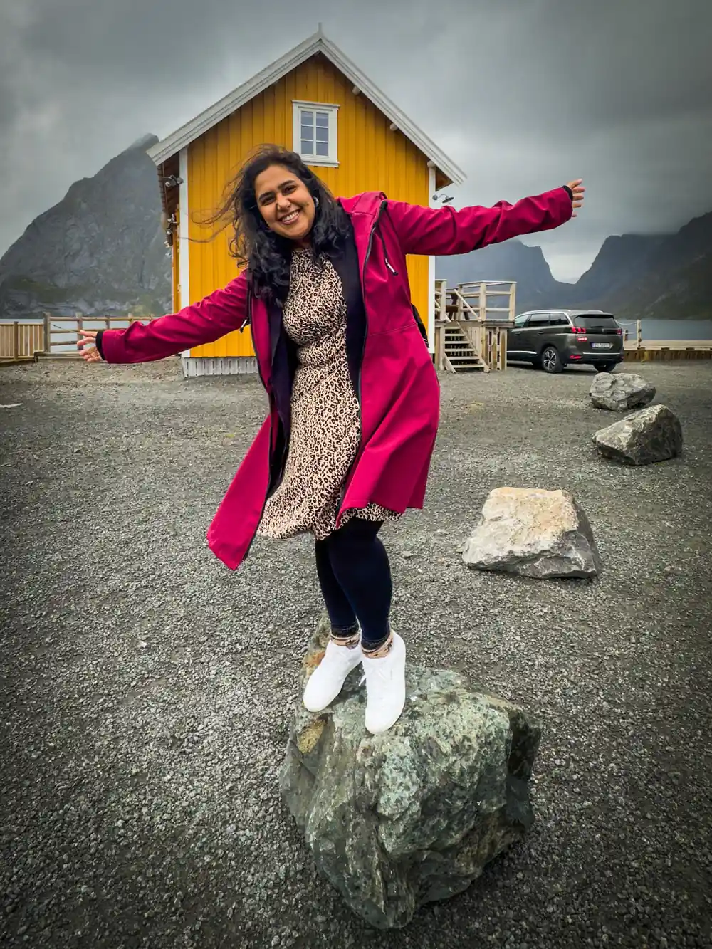 Woman in magenta jacket jumping on rock near yellow house